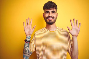 İzole edilmiş sarı arka plan üzerinde duran çizgili tişörtlü genç adam on numarayı gösterirken kendinden emin ve mutlu bir şekilde gülümsüyor..