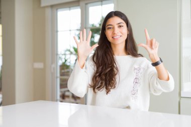 Genç güzel kadın evde beyaz masada gösteren ve parmakları ile işaret ederken yedi numara kendinden emin ve mutlu gülümseyerek.