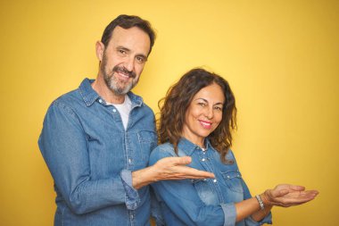 Güzel orta yaşlı çift, izole edilmiş sarı arka planda duruyorlar. Avuç içleri açık bir şekilde fotokopi alanını gösteriyorlar. Gülümseyerek mutlu bir şekilde gülümsüyorlar.