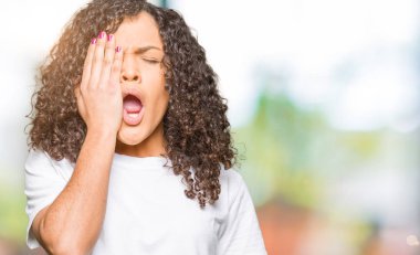 Kıvırcık saçlı genç güzel kadın beyaz t-shirt giyen Yaslama yorgun yarım yüz kapsayan, göz ve el ile ağız. Yüz acı içinde acıyor.