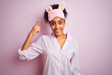 Genç Afro-Amerikalı kadın pijama giyiyor ve maske takıyor, pembe arka planda, tebessüm ediyor tek parmağıyla, harika bir fikir ya da düşünceyle, iyi bir hafıza.