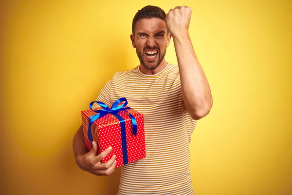 Young man holding a birthday present over yellow isolated background annoyed and frustrated shouting with anger, crazy and yelling with raised hand, anger concept
