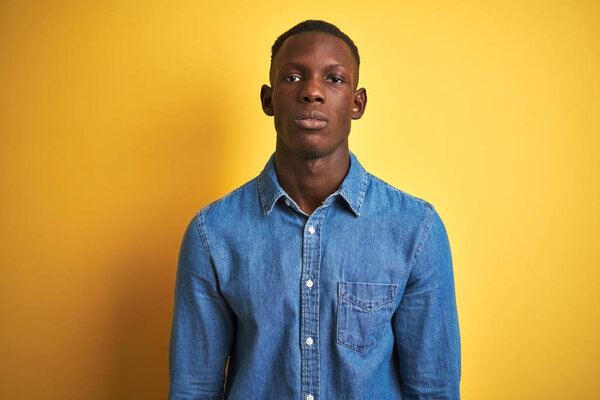 Young african american man wearing denim shirt standing over isolated yellow background with serious expression on face. Simple and natural looking at the camera.