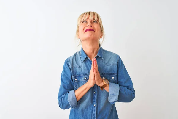Middle age woman wearing casual denim shirt standing over isolated white background begging and praying with hands together with hope expression on face very emotional and worried. Asking for forgiveness. Religion concept.