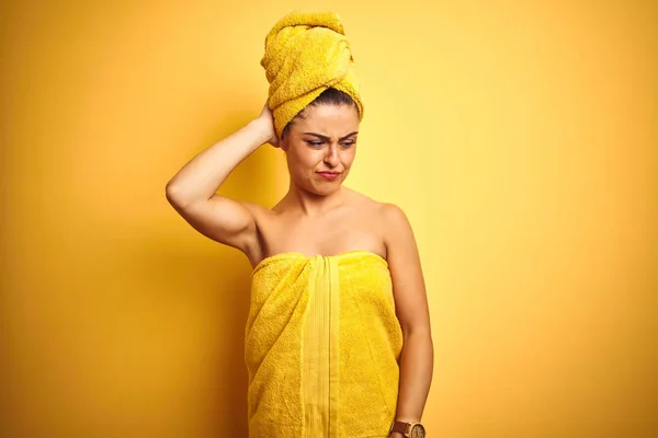 Young beautiful woman wearing towel after shower over isolated yellow background confuse and wonder about question. Uncertain with doubt, thinking with hand on head. Pensive concept.