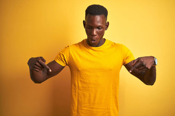Young african american man wearing casual t-shirt standing over isolated yellow background Pointing down with fingers showing advertisement, surprised face and open mouth