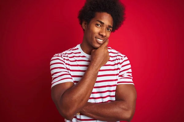 アフリカ系アメリカ人の若い男性が 赤い背景の上にストライプのTシャツを着ていて カメラが交差する腕とあごに上げられた手で笑顔で自信を持っているのを見ている 肯定的な考え — ストック写真