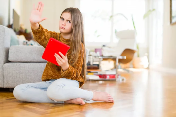 Beautiful young girl kid using digital touchpad tablet sitting on the floor with open hand doing stop sign with serious and confident expression, defense gesture