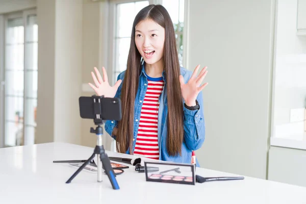 Beautiful Asian influencer woman recording make up tutorial using smartphone camera crazy and mad shouting and yelling with aggressive expression and arms raised. Frustration concept.