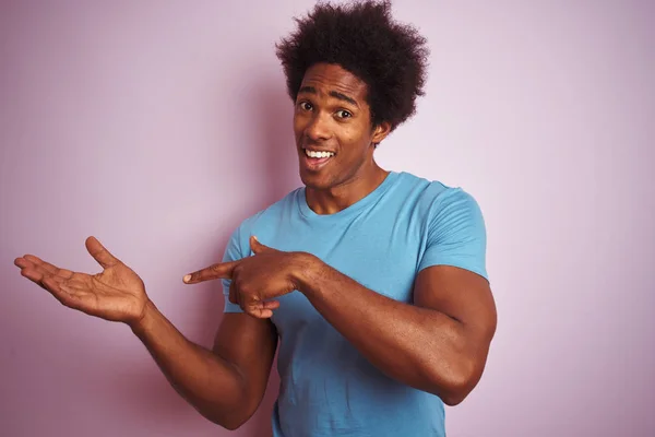 African american man with afro hair wearing blue t-shirt standing over isolated pink background amazed and smiling to the camera while presenting with hand and pointing with finger.