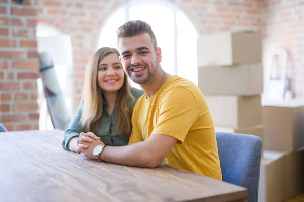 年轻漂亮的夫妇坐在家里的桌子上 拥抱在爱很高兴搬进新家 他们身后的纸板箱 — 图库照片