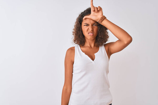 Young brazilian woman wearing casual t-shirt standing over isolated white background making fun of people with fingers on forehead doing loser gesture mocking and insulting.