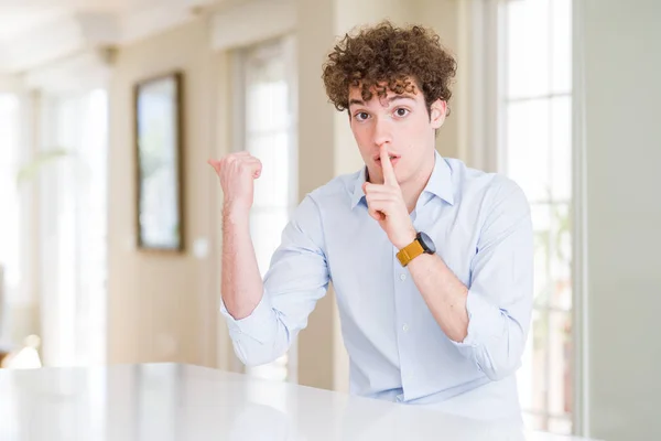 Young business man with curly read head asking to be quiet with finger on lips pointing with hand to the side. Silence and secret concept.