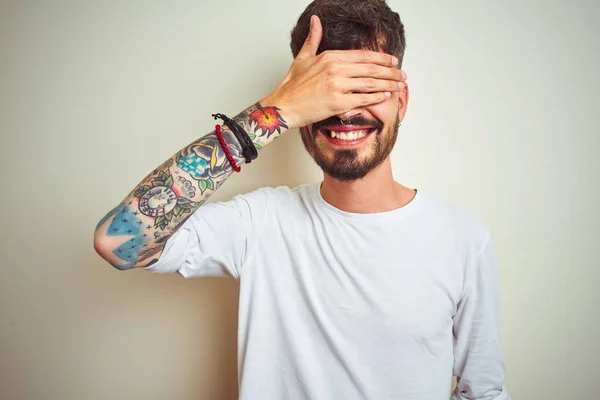 Young man with tattoo wearing t-shirt standing over isolated white background smiling and laughing with hand on face covering eyes for surprise. Blind concept.