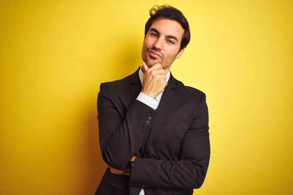 Young handsome businessman wearing suit and tie standing over isolated yellow background looking confident at the camera smiling with crossed arms and hand raised on chin. Thinking positive.