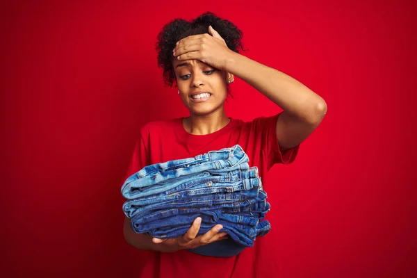 Young african american woman holding stack of jeans over isolated red background stressed with hand on head, shocked with shame and surprise face, angry and frustrated. Fear and upset for mistake.