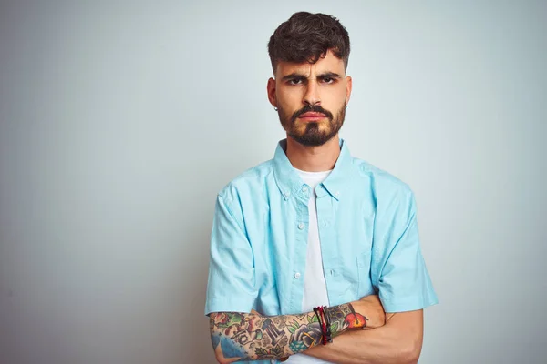 Young man with tattoo wearing blue shirt standing over isolated white background skeptic and nervous, disapproving expression on face with crossed arms. Negative person.