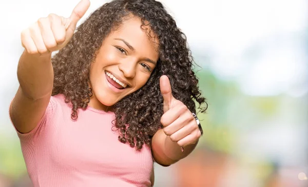 Young beautiful woman with curly hair wearing pink t-shirt approving doing positive gesture with hand, thumbs up smiling and happy for success. Looking at the camera, winner gesture.