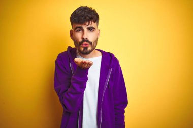 İzole edilmiş sarı arka plan üzerine spor mor kazak giyen genç bir adam havayla öpüşürken kameraya bakıyor. Güzel ve seksi. Aşk ifadesi.