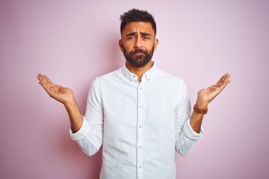 Genç Hintli iş adamı zarif bir gömlek giyerek soyutlanmış pembe arka plan üzerinde duruyor. Kolları ve elleri havada, kafası karışmış bir ifade takınıyor. Şüphe kavramı.