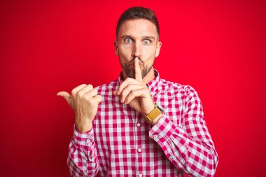 Kırmızı izole edilmiş arka plan üzerinde genç yakışıklı bir adam sessiz olmak istiyor. Parmakları dudaklarında, elleri yandan işaret ederek. Sessizlik ve gizli konsept.
