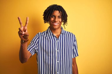 Rastgele çizgili tişörtler giyen Afro adam izole edilmiş sarı arka planda iki numarayı gösterirken kendinden emin ve mutlu bir şekilde gülümsüyor..