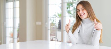 Wide angle picture of beautiful young girl kid wearing casual sweater approving doing positive gesture with hand, thumbs up smiling and happy for success. Looking at the camera, winner gesture. clipart