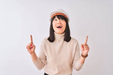 Genç, güzel, miğfer takan Çinli bir mimar beyaz arka planda şaşkın şaşkın yukarı bakıyor, parmaklarıyla işaret ediyor ve kollarını kaldırıyordu..