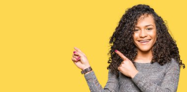 Kıvırcık saçlı giyen ile genç güzel gri kazak gülümseyen kadın ve iki elleri ve parmakları ile tarafına işaret kamera bakıyor.