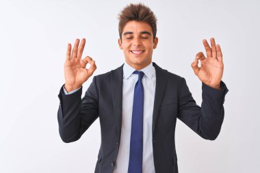 İzole edilmiş beyaz arka plan üzerinde duran takım elbiseli genç yakışıklı işadamı rahatla ve parmaklarıyla meditasyon hareketi yaparken gözleri kapalı gülümse. Yoga konsepti.