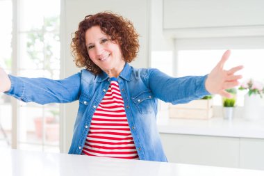 Kıvırcık saçlı, kot ceket giyen orta yaşlı bir kadın kameraya sarılıp gülümsüyor. Mutluluğu kucaklayan neşeli bir ifade..