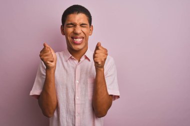 İzole edilmiş pembe arka planda dikilen, kolları havada ve gözleri kapalı zaferi kutlayan genç, yakışıklı Arap adam. Kazanan konsept.