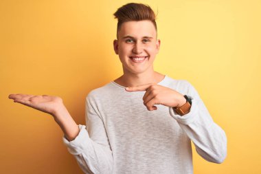 İzole edilmiş sarı arka plan üzerinde duran beyaz tişörtlü genç yakışıklı adam kameraya hayret ediyor ve gülümsüyor..