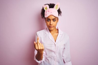 Genç Afro-Amerikalı kadın pijama giyiyor ve maske takıyor pembe arka planda, orta parmağını gösteriyor, kaba ve kaba bir ifade.