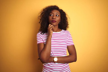 Genç Afro-Amerikalı kadın izole edilmiş sarı arka plan üzerinde çizgili tişört giyiyor ve elleri çenesinde soru sormayı, düşünceli ifadeyi düşünüyor. Düşünceli bir yüzle gülümsüyor. Şüphe kavramı.