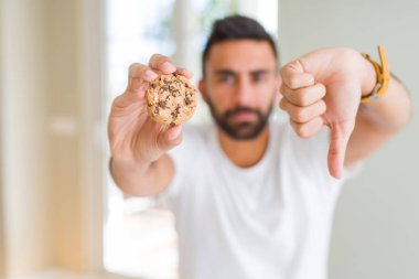 Yakışıklı İspanyol adam kızgın yüz ile çikolata parçaları kurabiye yeme, başparmak aşağı, ret kavramı ile sevmediğimi gösteren negatif işareti