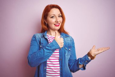 Güzel kızıl saçlı kadın kot gömlek ve çizgili tişört giyerek izole pembe arka plan üzerinde avuç içini gösteriyor ve baş parmaklarıyla iyi bir hareket yapıyor, mutlu ve neşeli gülümsüyor.