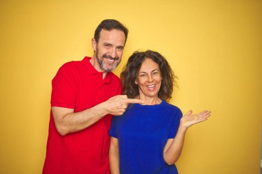 Güzel orta yaşlı çift, izole edilmiş sarı arka planda durarak hayretler içinde kameraya bakıp gülümserken elleri ve parmaklarıyla işaret ediyorlar..