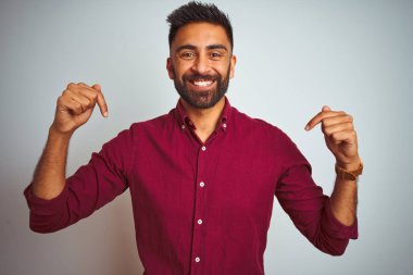 İzole edilmiş gri arka planda duran kırmızı şık gömlekli genç Hintli adam yüzünde bir gülümsemeyle gururlu ve mutlu birini işaret ediyor..