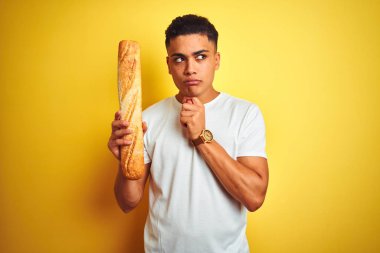 Genç Brezilyalı fırıncı, izole edilmiş sarı arka planda duran ekmeği tutuyor. Ciddi bir yüz ifadesi var. Soru sormayı düşünüyor.