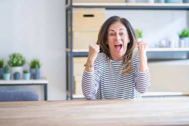Evde masada oturan orta yaş kıdemli kadın şaşırttı ve silah kaldırdı ve açık gözleri ile başarı için hayran kutluyor. Kazanan kavramı.