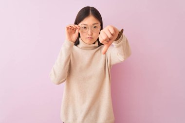 Balıkçı yaka kazak ve gözlük takan genç Çinli bir kadın. İzole edilmiş pembe arka plan, kızgın yüz, negatif işaret baş parmakları aşağıda, reddedilme konsepti.