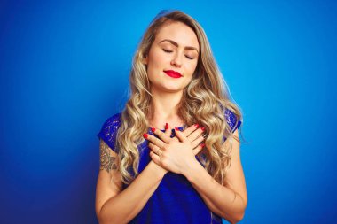 Mavi izole edilmiş arka planda duran genç ve güzel bir kadın elleri göğsünde, gözleri kapalı ve yüzünde minnet dolu bir jest ile gülümsüyor. Sağlık kavramı.