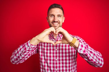 Kırmızı izole edilmiş arka plan üzerinde genç yakışıklı bir adam. Sevgiyle gülümsüyor. Elleriyle kalp sembolü yapıyor. Romantik konsept.