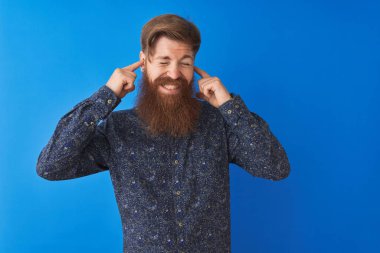 Çiçekli yaz tişörtü giyen genç kızıl saçlı adam izole edilmiş mavi arka planda duruyor. Yüksek sesli müzik sesi için parmaklarıyla kulaklarını kapatıyor. Sağır kavramı.