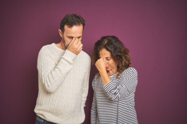 Güzel bir orta yaşlı çift, izole edilmiş mor arka plan üzerine kazak giyip yorgun burunlarını ve gözlerini ovuşturup yorgunluk ve baş ağrısı hissediyorlar. Stres ve hüsran kavramı.