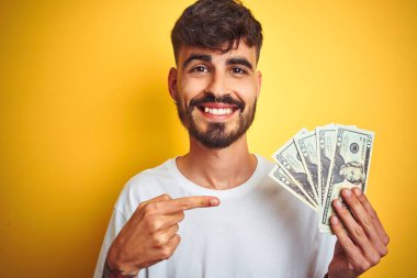Dövmeli genç adam, izole edilmiş sarı arka planda dolarlar tutuyor. Elle ve parmakla işaret ederek çok mutlu.