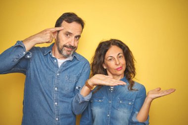 Güzel orta yaşlı çift, izole edilmiş sarı arka planda dikiliyorlar. Avuç içleri açık, fotokopi alanı gösteriliyor ve alnını işaret ediyorlar. Bunu bir düşün..