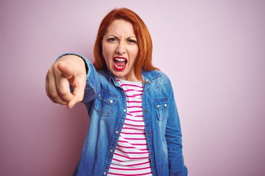 Güzel kızıl saçlı kadın, kot gömlek ve çizgili tişört giyip pembe arka planda izole edilmiş. Kameraya kızgın ve sinirli olduğunu gösteriyor.