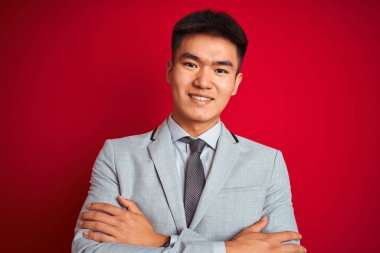 Gri ceketli ve kravatlı Asyalı Çinli işadamı izole edilmiş kırmızı arka planda durup kameraya bakarken kollarını kavuşturup gülümsüyor. Pozitif kişi.
